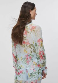 Femme aux longs cheveux bruns de profil, portant un chemisier bleu clair transparent et un pantalon assorti avec un motif floral rose et jaune.