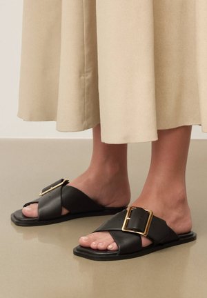 Noge v črnih usnjenih natikačih s širokimi križajočimi se trakovi in velikimi zlatimi pravokotnimi zaponkami, stojijo na sijajni tleh.