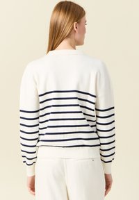 Weißer Strickpullover mit marineblauen horizontalen Streifen auf dem Rücken. Hat einen lockeren Schnitt, gerippte Bündchen und einen runden Halsausschnitt.