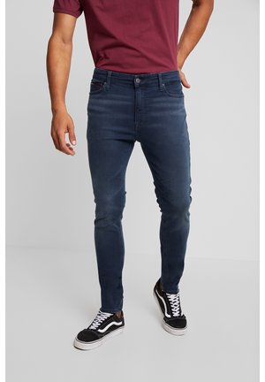 Man draagt slim-fit donkerblauwe jeans, een bordeaux rood t-shirt en zwart-witte sneakers, staand tegen een effen achtergrond.