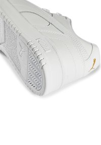 Sneaker en cuir blanc avec des accents texturés, semelle en caoutchouc et logo doré. Présente un design imposant et des détails de couture visibles.