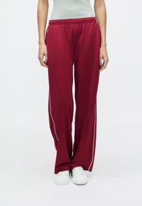 Vyno raudonos sportinės kelnės su laisvo kirpimo fitu, baltais šonais dryžiais, elastine juosmens dalimi, derinamos su baltais sportbačiais.