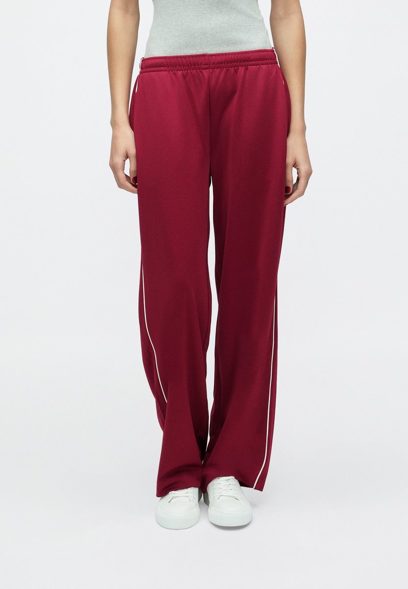 Vyno raudonos sportinės kelnės su laisvo kirpimo fitu, baltais šonais dryžiais, elastine juosmens dalimi, derinamos su baltais sportbačiais.
