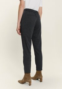 Frau, die dunkelgraue karierte, taillierte Hose mit elastischem Bund und braunen Wildleder-Stiefeletten trägt, von hinten vor einem hellen Hintergrund gezeigt.