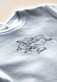 Un body en coton bleu clair avec un design brodé noir de personnages animaliers, accompagné du texte "Bambi" en dessous. Texture douce, encolure arrondie.