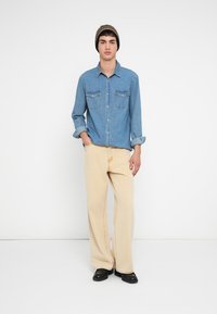 Camisa vaquera azul claro, con botones al frente y dos bolsillos en el pecho; combinada con pantalones anchos beige; zapatos negros; gorro de punto con detalles marrones.