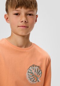 Koszulka z krótkim rękawem, okrągłym dekoltem w jasnopomarańczowym kolorze z zielonym haftowanym wzorem liścia na klatce piersiowej. Gładka tekstura, luźny krój.