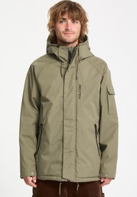 Veste imperméable vert olive avec capuche, devant zippé, poches latérales et logo Volcom. Présente une coupe décontractée et un tissu texturé.