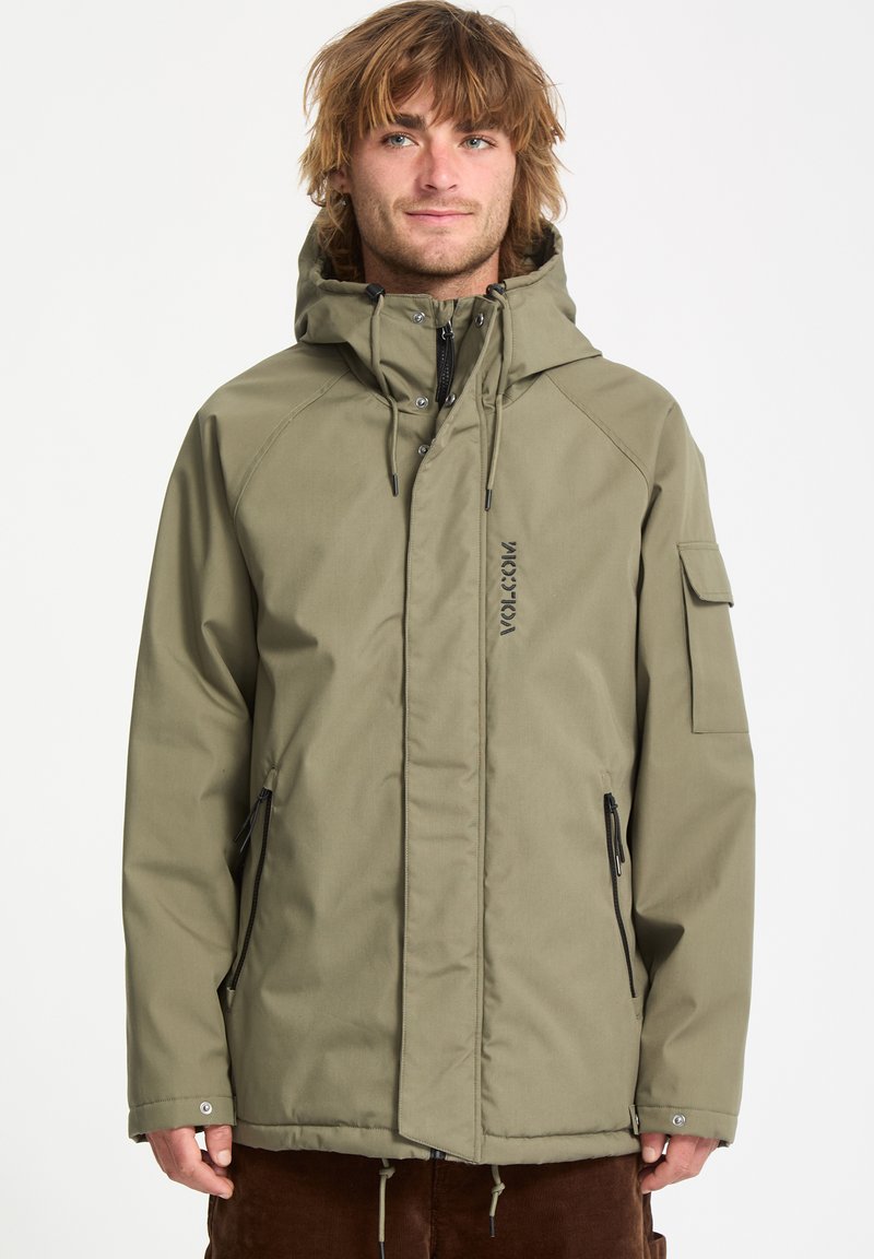 Veste imperméable vert olive avec capuche, devant zippé, poches latérales et logo Volcom. Présente une coupe décontractée et un tissu texturé.