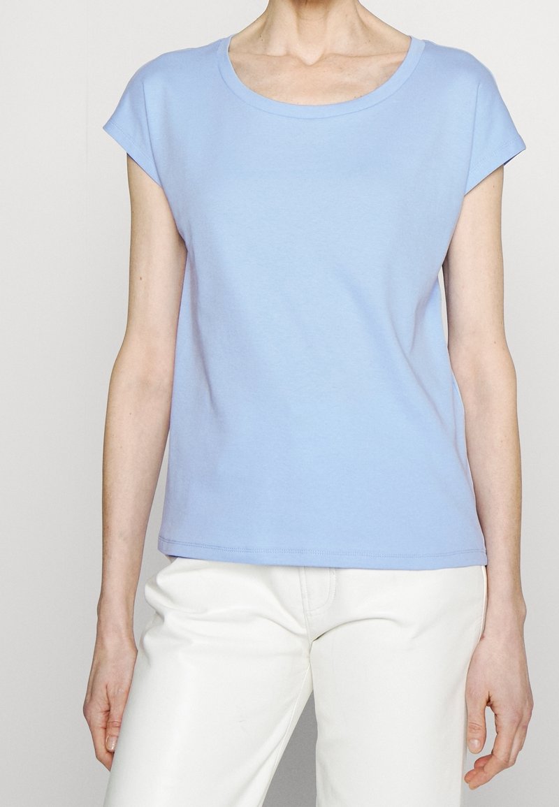Personne portant un haut bleu clair à manches courtes et un pantalon blanc, se tenant devant un fond clair uni.