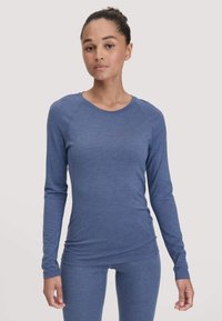 Jonge vrouw in een strak blauw longsleeve top en bijpassende leggings, staand tegen een effen lichte achtergrond.