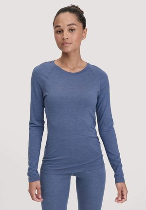 Giovane donna che indossa un top a maniche lunghe blu aderente e leggings abbinati, in piedi contro uno sfondo chiaro e semplice.
