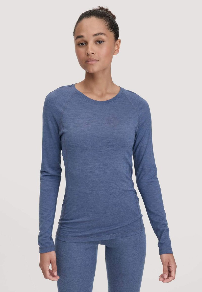 Jonge vrouw in een strak blauw longsleeve top en bijpassende leggings, staand tegen een effen lichte achtergrond.