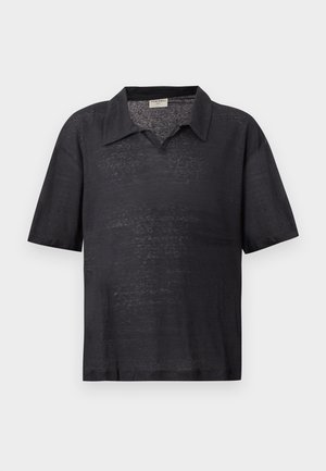 Άνδρας με σγουρά μαλλιά που φοράει μαύρο κοντομάνικο πουκάμισο με γιακά, μπεζ φαρδύ παντελόνι και μαύρα σανδάλια, στέκεται με τα χέρια στις τσέπες.