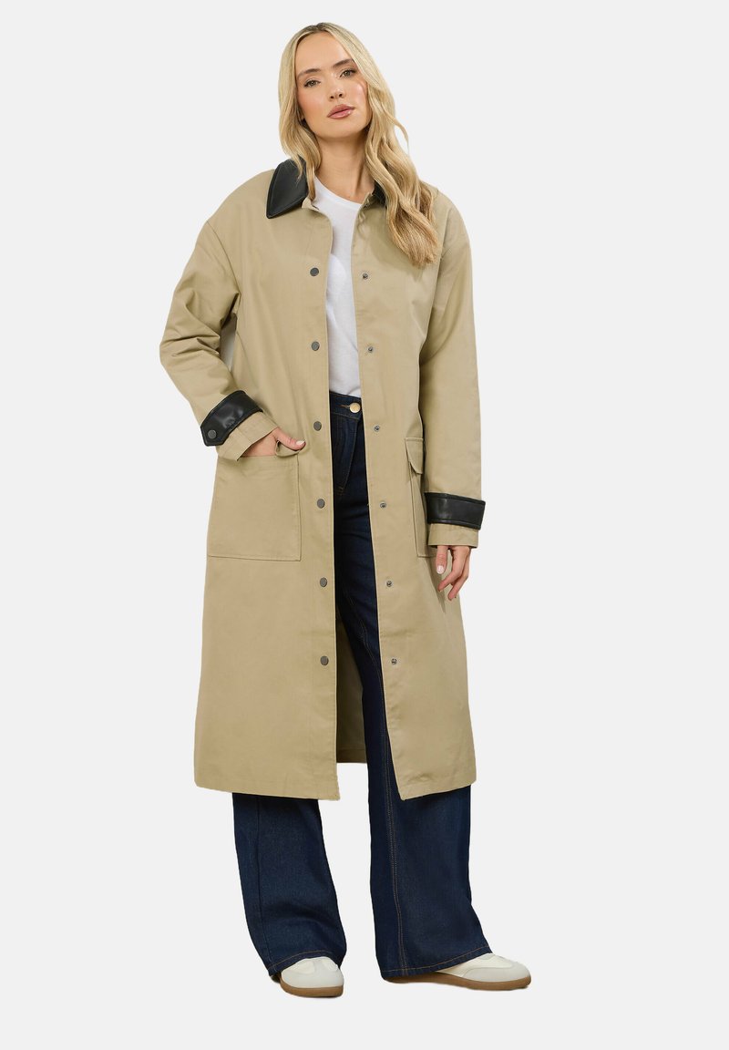 Cappotto lungo beige con colletto e polsini neri, dotato di bottoni a scatto, tasche laterali e una vestibilità dritta, indossato sopra jeans a gamba larga blu.