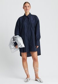 Marineblaues Oversize-Hemd aus leichtem Stoff, mit klassischem Kragen, langen Ärmeln und einer silbernen metallischen Tasche in der Hand. Silberne flache Schuhe mit überkreuzten Riemen.