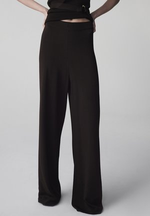 Persona che indossa pantaloni neri a gamba larga, larghi e a vita alta e un top nero abbinato con fibbia alla cintura su uno sfondo chiaro uniforme.