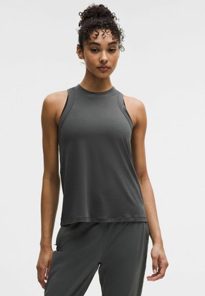 Femme aux cheveux bouclés portant un haut sans manches gris foncé et un pantalon assorti, debout devant un fond clair uni.