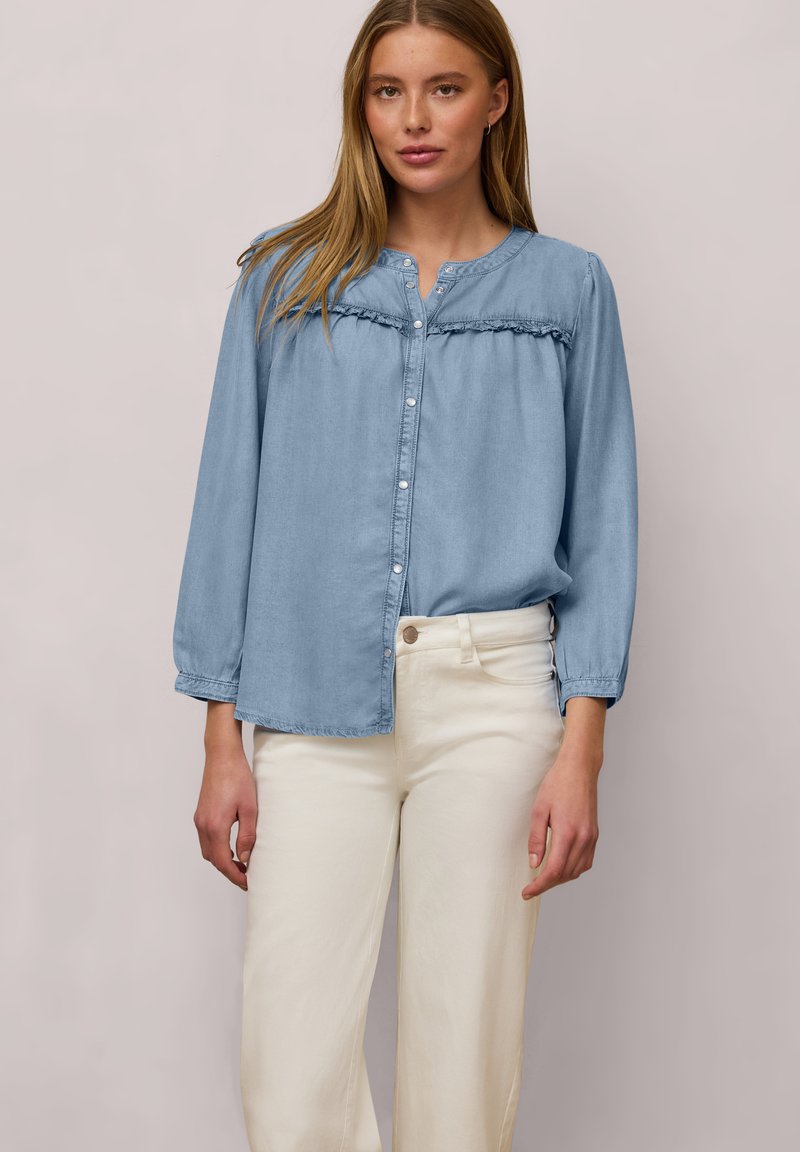 Femme portant un chemisier bleu clair à boutons avec des détails de volants et des manches longues, associé à un pantalon taille haute blanc cassé.