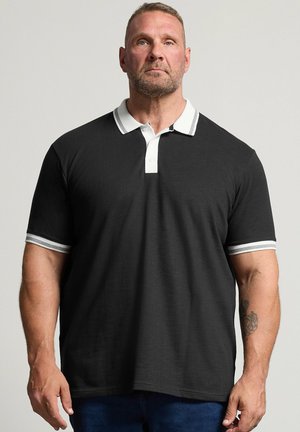 Mand med kort hår og skæg iført en sort polo-shirt med hvide krave- og manchetstriber, stående mod en lys baggrund.