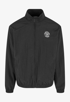 Schwarze Windjacke, mit Reißverschluss, hohem Kragen, elastischen Bündchen und Saum. Mit einem runden Logo auf der linken Brust. Leichtes, glattes Material.