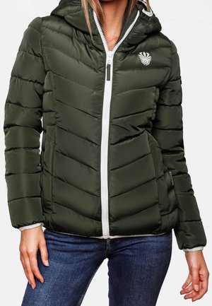 Femme portant une veste matelassée vert foncé avec une fermeture éclair blanche et un logo sur la poitrine, associée à un jean bleu, sur un fond uni.