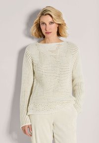 Blonde Frau mit grünen Augen, die einen cremefarbenen, strukturierten Strickpullover und cremefarbene Ripp-Hosen trägt, steht vor einem hellen Hintergrund.