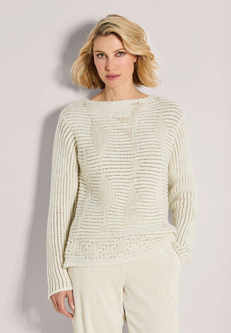 Blonde Frau mit grünen Augen, die einen cremefarbenen, strukturierten Strickpullover und cremefarbene Ripp-Hosen trägt, steht vor einem hellen Hintergrund.