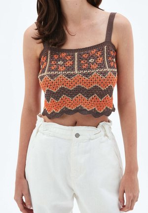 Femme portant un crop top crocheté marron et orange avec un motif en zigzag et un pantalon blanc taille haute sur un fond uni.