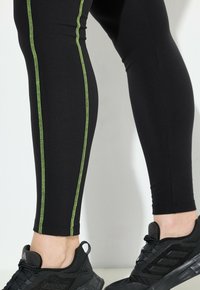 Leggings negros con costuras verdes contrastantes a lo largo de los lados, con un diseño ajustado y combinados con zapatillas deportivas negras.