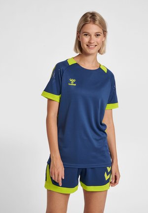 Młoda kobieta uśmiechnięta, ubrana w niebieską i neonowo zieloną sportową koszulkę oraz pasujące krótkie spodenki na jednolitym tle.