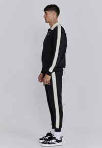 Survêtement noir avec des rayures blanches sur les côtés, veste zippée et pantalon de jogging. Présente un logo sur la poitrine et un tissu texturé. Baskets noires.