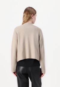 Beige avkortet genser med en myk tekstur, med lange ermer og rund hals, brukt over en svart skjorte og kombinert med svarte bukser.