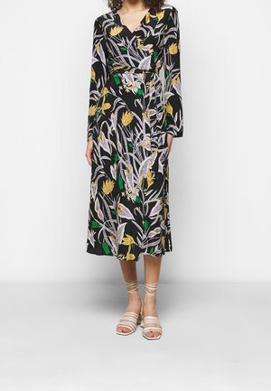 Kvinde iført en sort blomsterprint slå-om kjole med gule, hvide og grønne blade, kombineret med beige sandaler med stropper, stående mod en hvid baggrund.