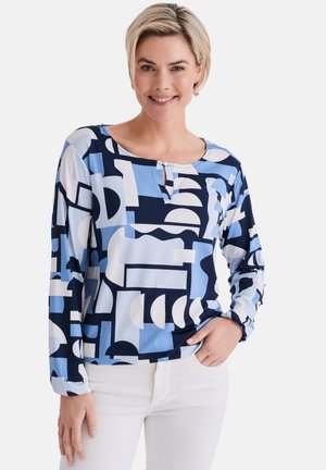 Lächelnde Frau, die ein langärmliges Oberteil mit blauem, marineblauem und weißem geometrischem Muster sowie weiße Hosen trägt, steht mit einer Hand in der Tasche.