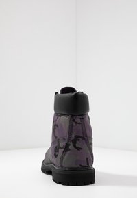 Botín de tobillo en camuflaje púrpura y negro con una superficie texturizada, suela de goma y cuello acolchado negro; costuras reforzadas en las uniones.