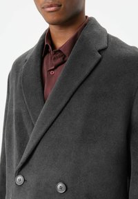 Dunkelgrauer, doppelt geknöpfter Mantel mit weicher Textur, Revers mit Kerbe und zwei schwarzen Knöpfen, kombiniert mit einem burgunderroten Hemd darunter.