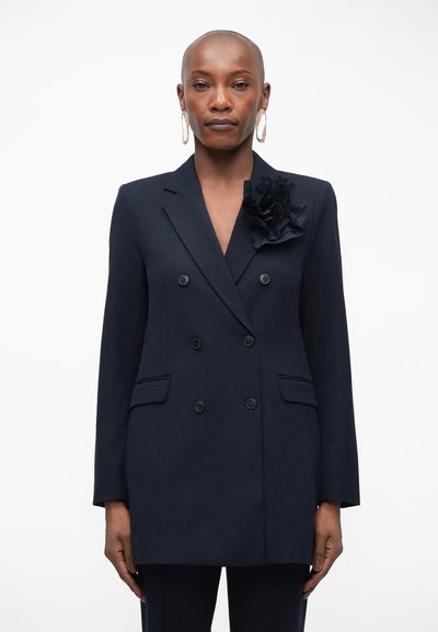 Blazer double boutonné bleu marine avec des revers crantés, deux poches à rabat, accent floral noir sur le côté gauche et texture de tissu lisse.