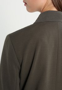 Blazer vert olive foncé avec un tissu texturé, un col épuré et un design sans couture, mettant en avant un style minimaliste et une coupe ajustée.
