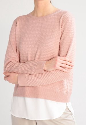 Mujer con un suéter rosa claro texturizado sobre una blusa blanca, con los brazos cruzados, de pie frente a un fondo claro y liso.