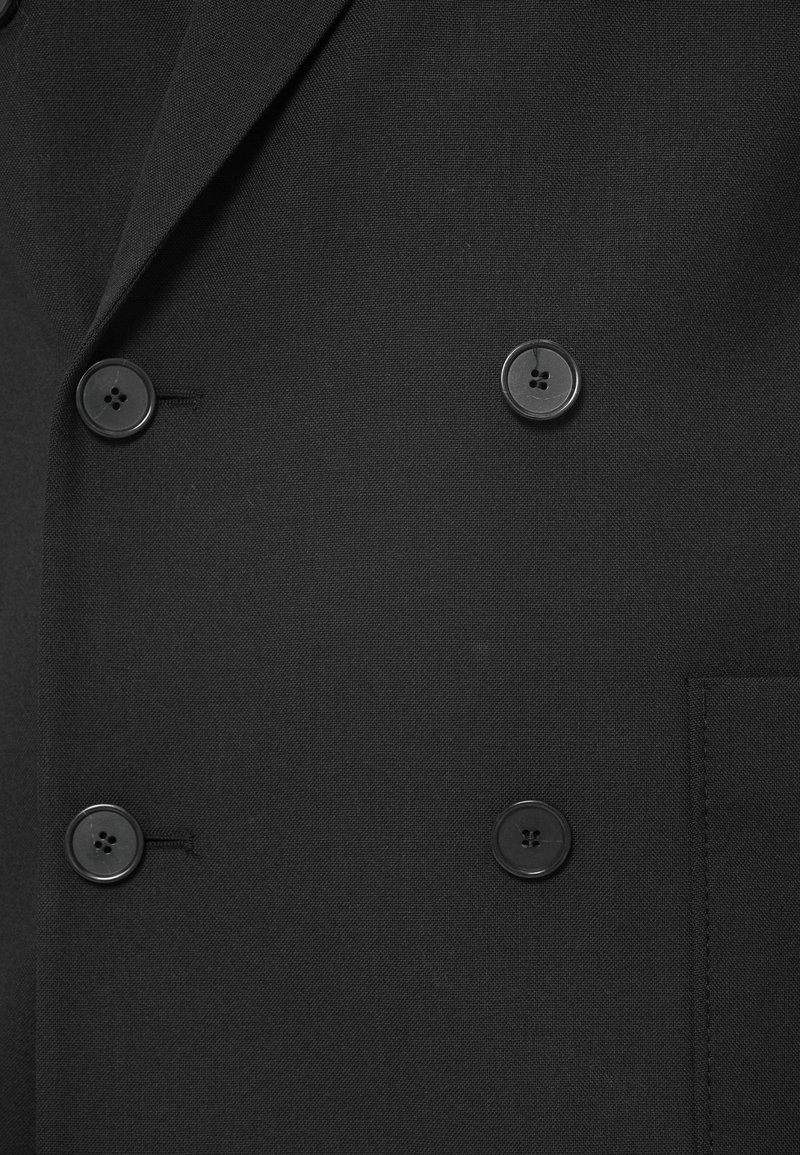 Sort dobbeltbrystet blazer med store, sorte knapper, der har en glat overflade og skarpe revers. Indeholder detaljer med sidelommer.