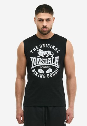 Uomo con capelli corti e barba indossa una maglietta da boxe nera senza maniche Lonsdale London e pantaloncini neri abbinati su uno sfondo semplice.