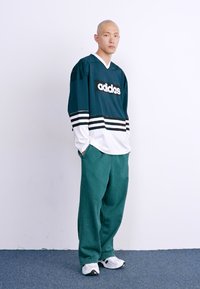 Zelen majica s črnimi in belimi črtami, z logotipom "adidas". V kombinaciji z ohlapnimi zelenimi hlačami in belimi tenisicami. Ozadje je enostavno.