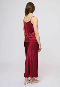 Camicia da notte in raso rosso con pizzo, caratterizzata da una gonna fluida e spalline regolabili, vista da dietro con scarpe beige senza chiusura.