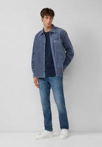 Camisa chaqueta de pana azul con un bolsillo en el pecho, usada sobre una camiseta de rayas azul marino, combinada con jeans de denim claros y zapatillas blancas.