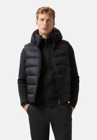 Młody mężczyzna w czarnej, ocieplanej kamizelce bez rękawów z kapturem, nosi pod nią zapięty czarny sweter i czarne spodnie, z rękami w kieszeniach.