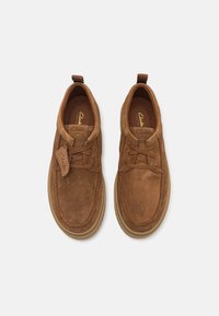 Brown suede casual shoes with laces, rounded toe, and rubber sole. Features a small branded tag near the lace on the side.