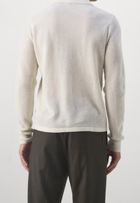 Maglione lavorato a maglia beige chiaro con un motivo testurizzato, polsini e orlo a coste, abbinato a pantaloni scuri, visto da dietro.