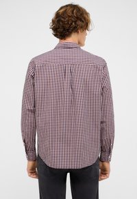 Mężczyzna o kręconych włosach, ubrany w długi rękaw w kratkę czerwono-białą i granatową oraz ciemne spodnie, pokazany plecami na jednolitym tle.