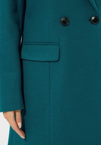 Cappotto in misto lana color teal con doppi bottoni, una tasca ben cucita e una finitura liscia e testurizzata sulle maniche.